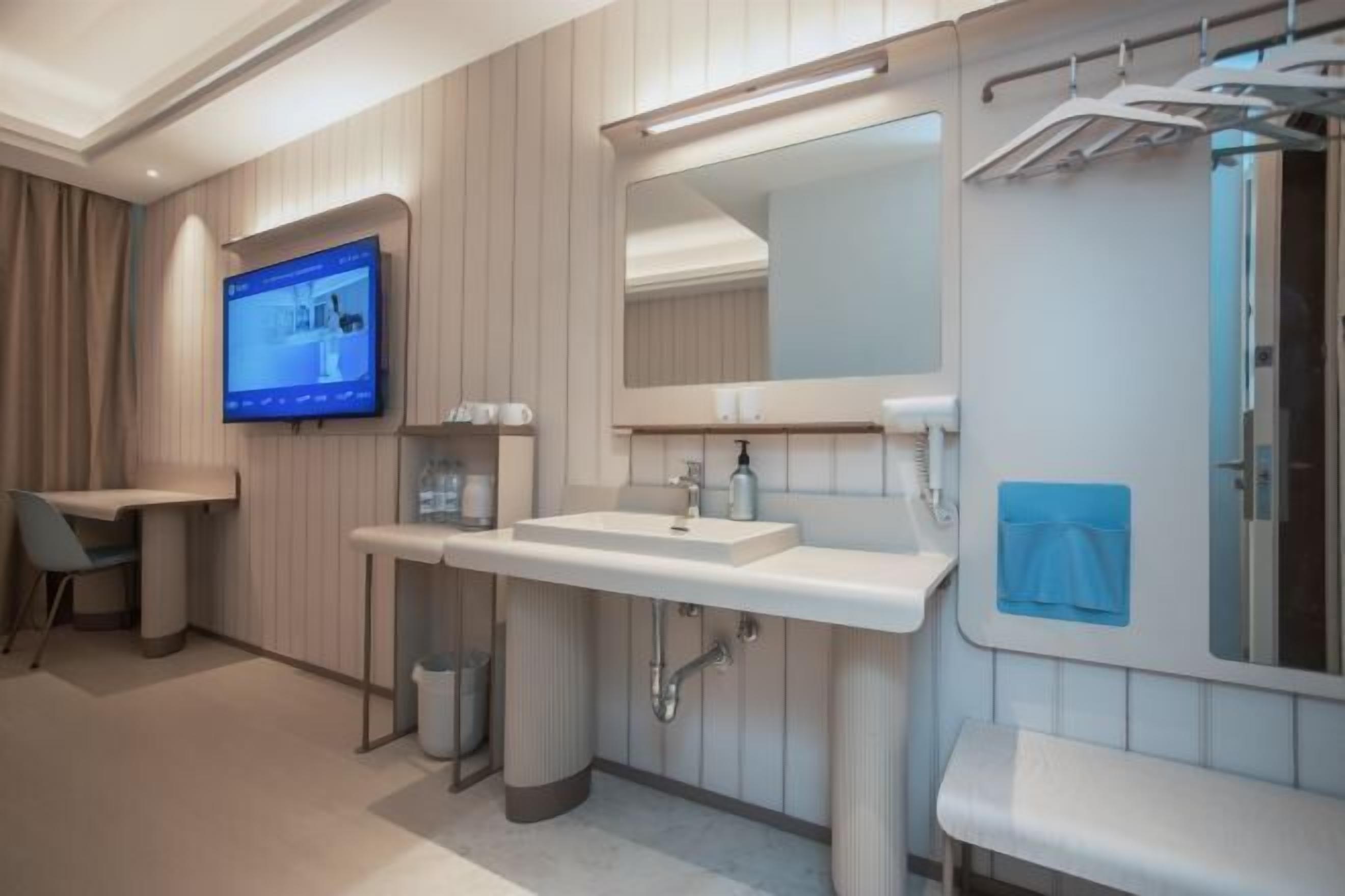 A modern bathroom with a sink, mirror, and shower area, including a TV and a trash can.