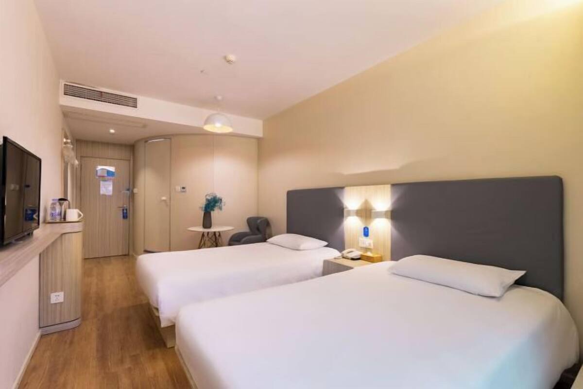 A hotel room with two beds, a desk, a chair, a TV, and a small table with a vase.