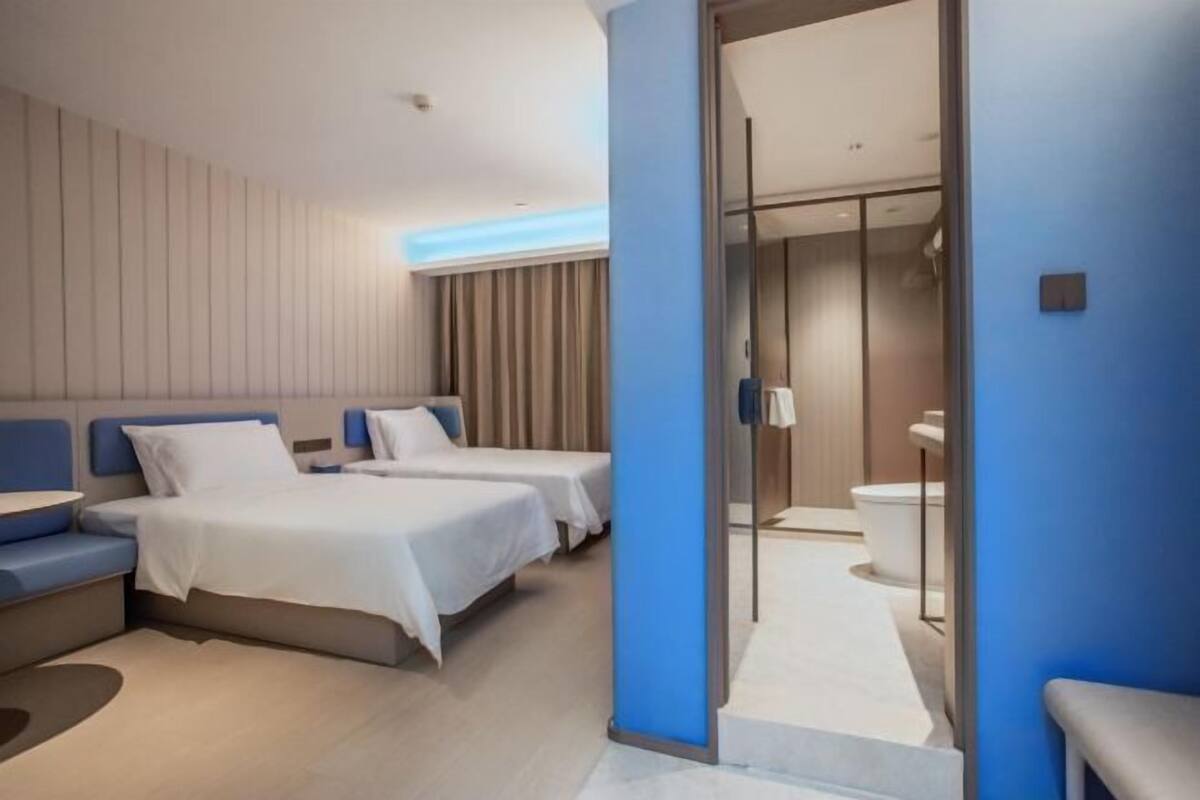 A modern hotel room with two beds, a seating area, and a bathroom with a glass partition.