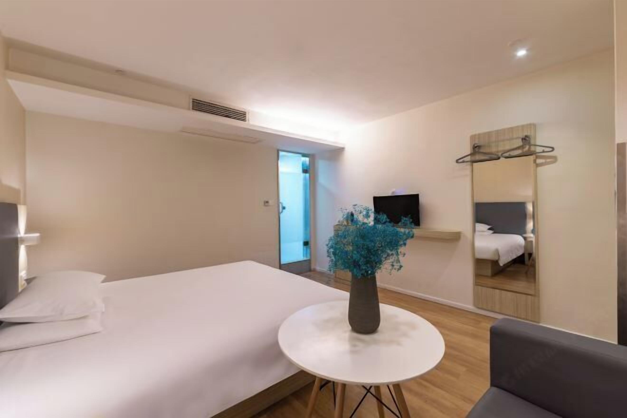 A modern hotel room with a bed, a round table with a vase, a TV, and a mirror.