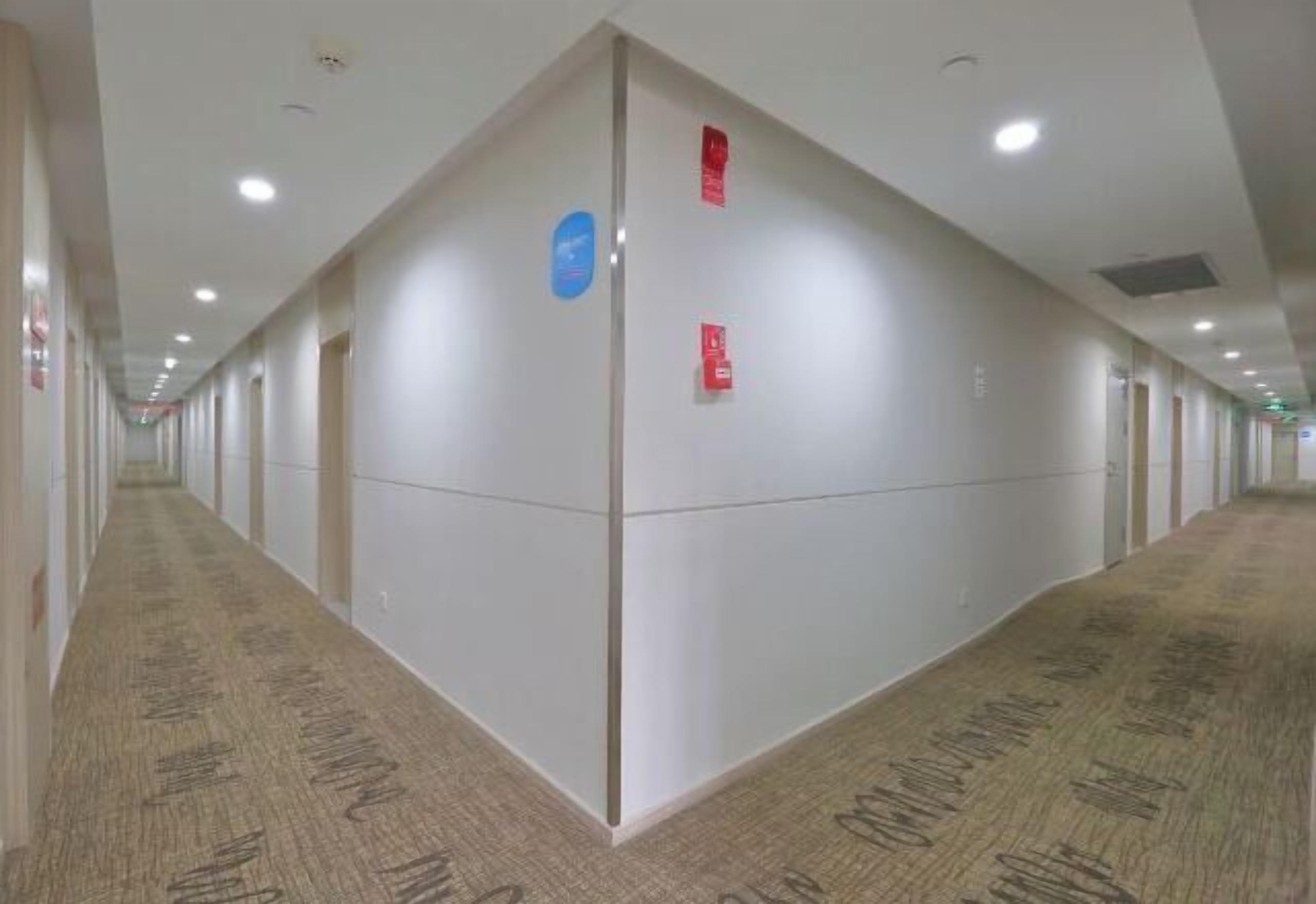 A long, narrow hotel corridor with a carpeted floor, white walls, and ceiling lights.