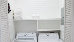 Un cuarto de lavado con dos lavadoras blancas de carga frontal y una secadora, piso de madera y una pared de azulejos blancos con un estante pequeño sobre las máquinas.