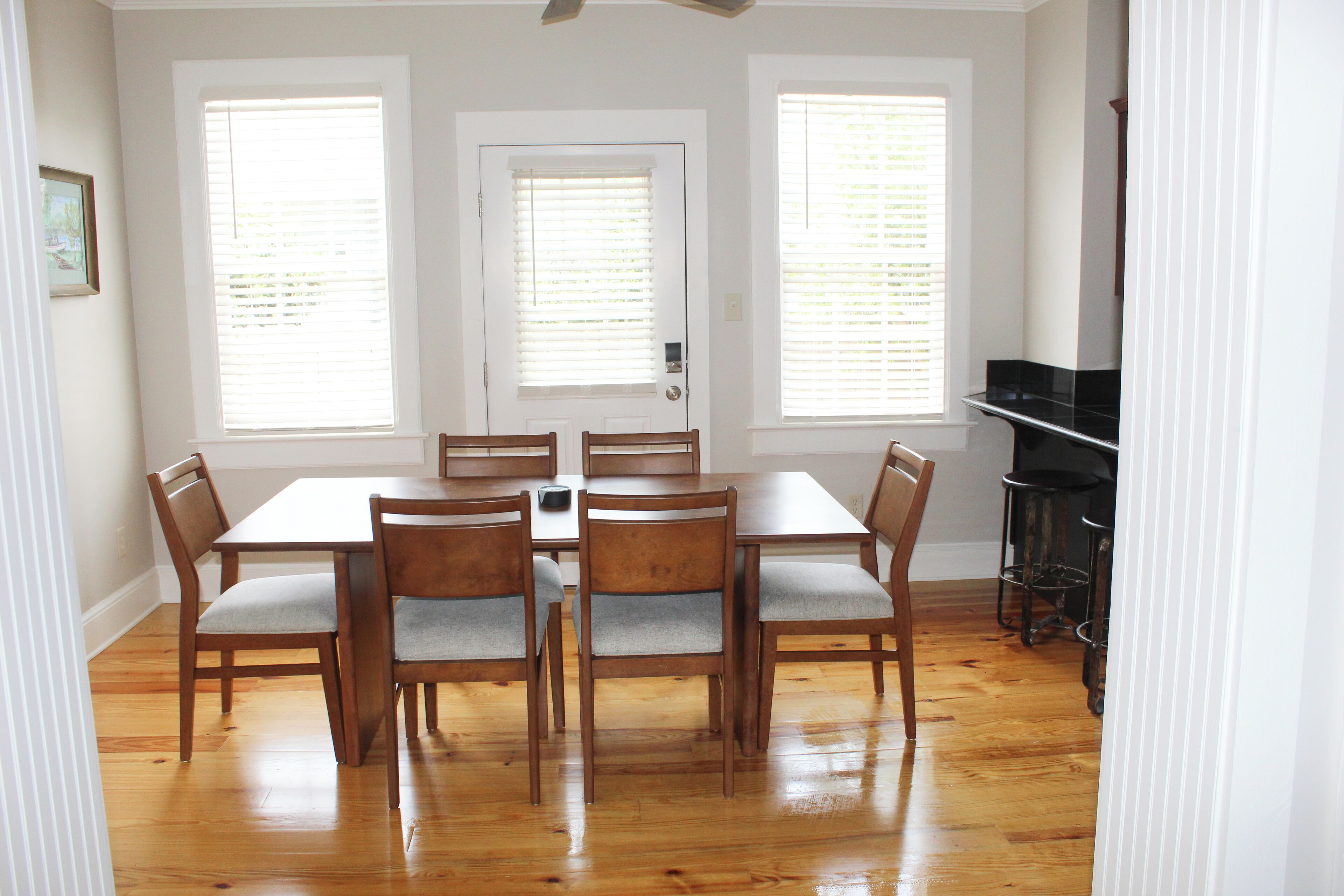 Uma sala de jantar com mesa de madeira e seis cadeiras, janelas amplas com persianas e uma porta.