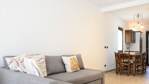 A modern living room with a grey sofa, a dining table with chairs, and a kitchen area visible in the background.