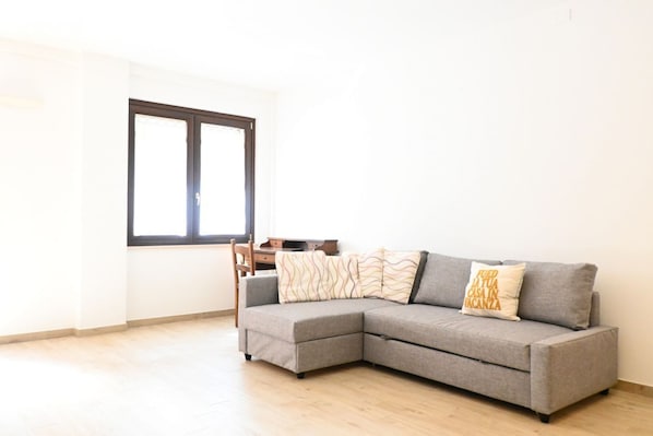 A grey sectional sofa with patterned cushions, a wooden chair, and a desk with a computer.