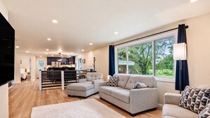 Ein modernes Wohnzimmer mit einem grauen Sofa, einem weißen Teppich und einem großen Fenster mit Vorhängen.