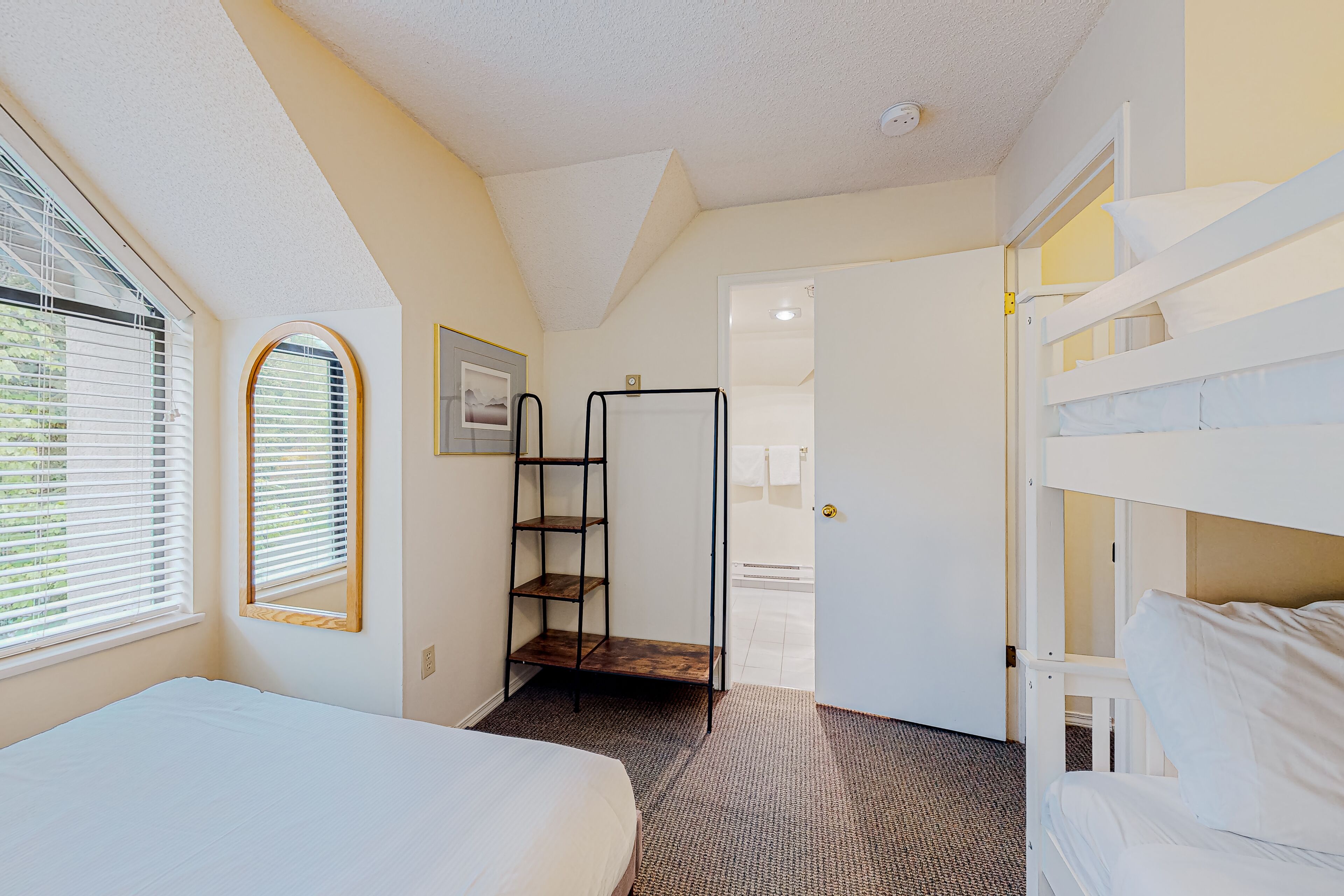 A bedroom with a bunk bed, a single bed, a wooden ladder shelf, a door leading to a bathroom, and a window with blinds.