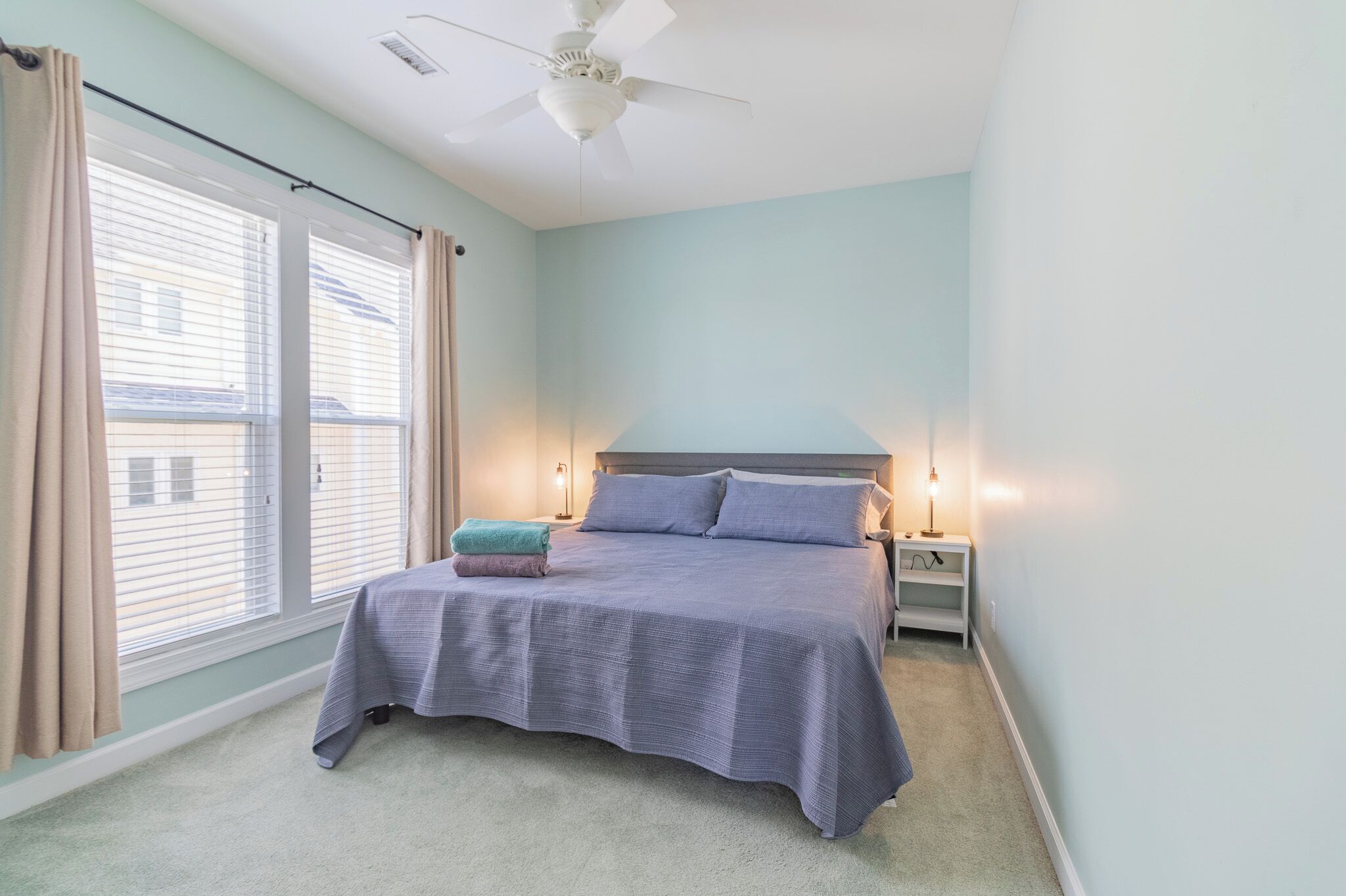 A bedroom with a bed, two bedside lamps, a nightstand, a ceiling fan, and a window with curtains.