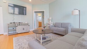 Ett vardagsrum med en grå soffa, ett runt soffbord, en tv som är monterad på väggen, ett skåp med hyllor och en lampa.