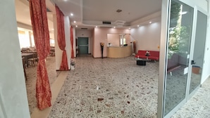 A spacious hotel lobby with a terrazzo floor, a reception desk, a red sofa, and a glass door leading outside. - hotel Anny  (Cervia)