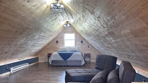 A spacious attic bedroom with a sloped ceiling, a large bed, a dark-colored sofa, and a wooden table. - Charming 2-bedroom cabin minutes from Sacandaga Lake and Saratoga Springs (Amsterdam)