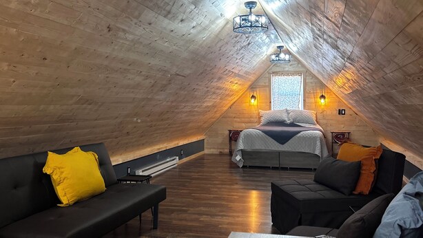 A cozy attic bedroom with wooden walls and a sloped ceiling. It features a bed, a sofa, and a dining table. - Charming 2-bedroom cabin minutes from Sacandaga Lake and Saratoga Springs (Amsterdam)