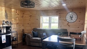 A cozy cabin interior with a wooden ceiling, a dining table with chairs, a sofa, and a kitchen area with a coffee maker and toaster. - Charming 2-bedroom cabin minutes from Sacandaga Lake and Saratoga Springs (Amsterdam)
