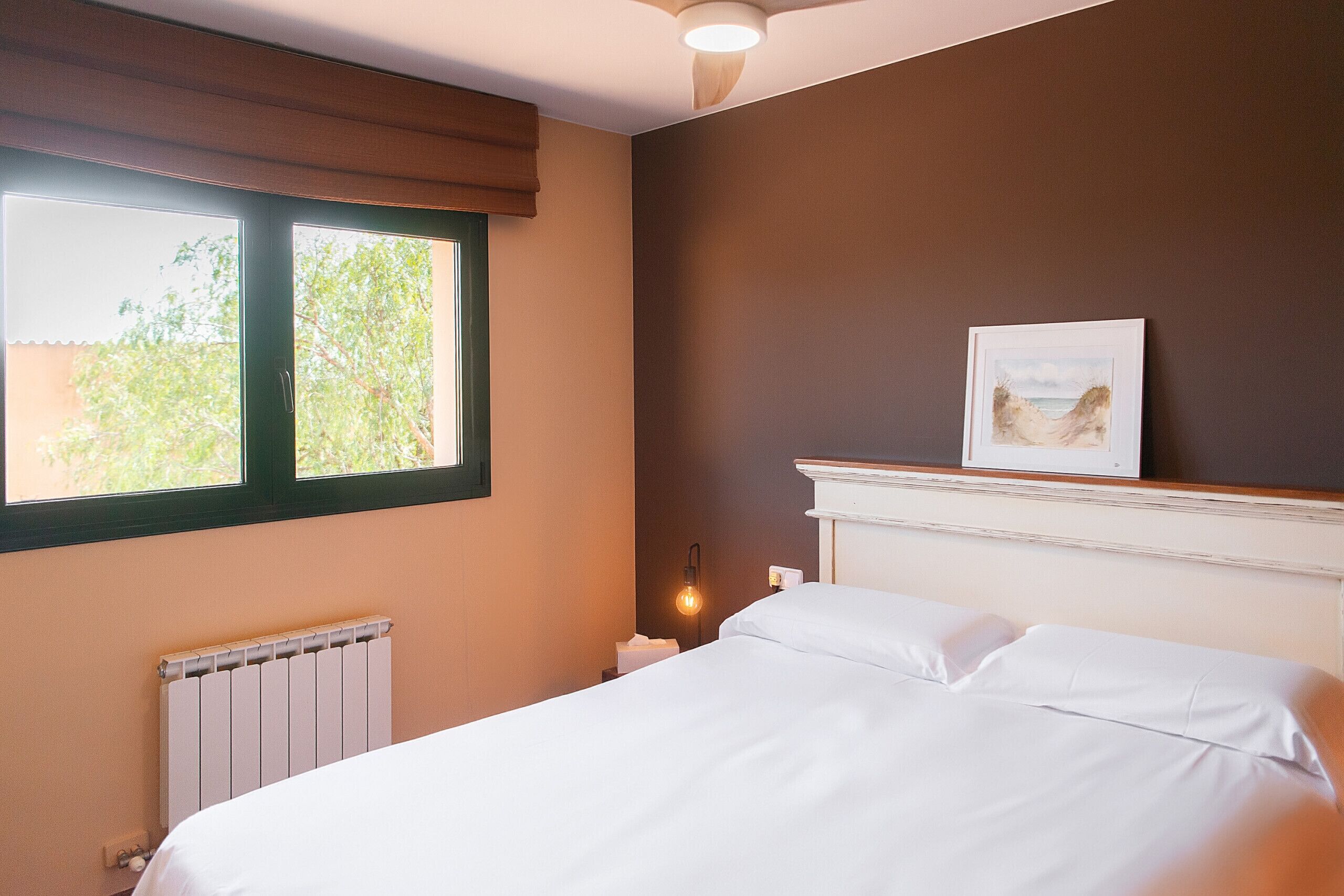 A hotel room with a bed, a radiator, a window with a view of trees, a wall-mounted picture, and a bedside table with a lamp.