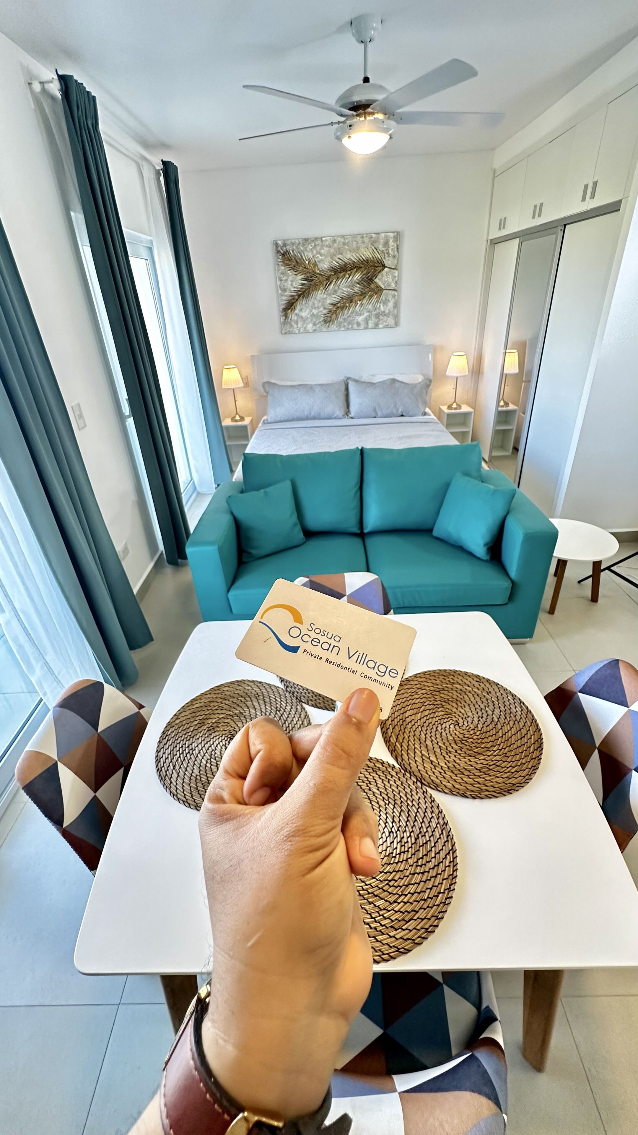 Ein Hotelzimmer mit einem Bett, einem türkisfarbenen Sofa, einem Esstisch mit Untersetzern und einem Deckenventilator.