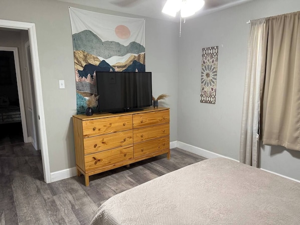 Une chambre avec une commode en bois, un téléviseur à écran plat, un lit recouvert d’une couverture beige, une tapisserie représentant un paysage de montagne et une décoration murale à motifs.