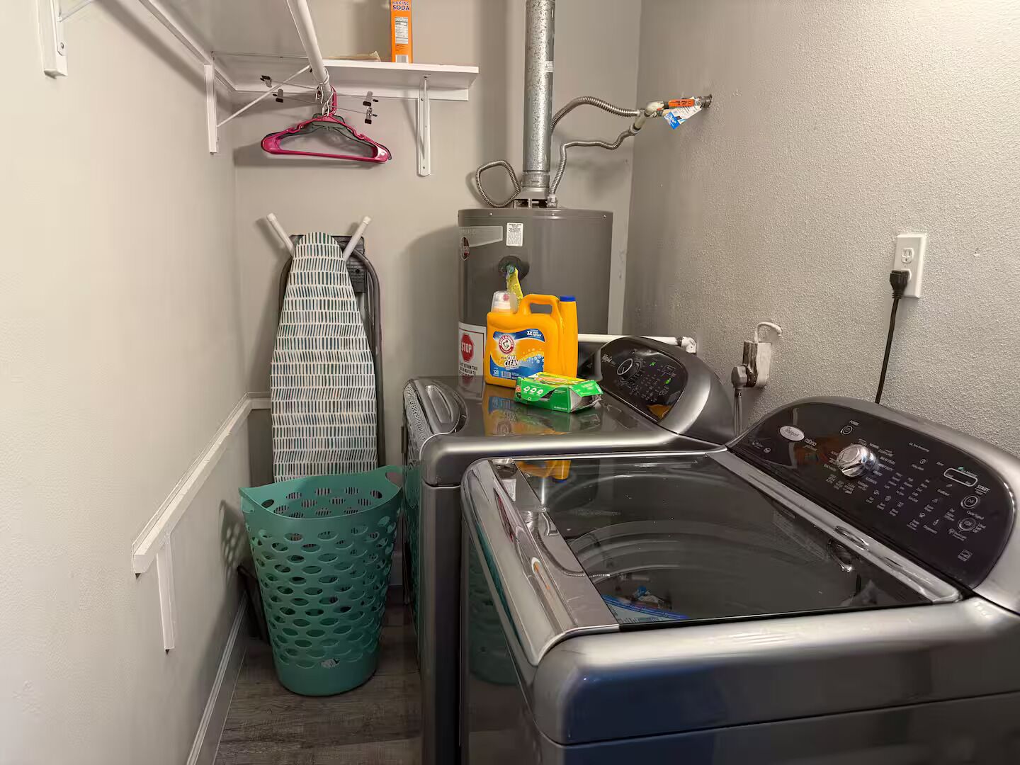 Ett tvättrum med en tvättmaskin med frontmatning, en torktumlare, en grön tvättkorg, en torkställning och en hylla med rengöringsmedel.