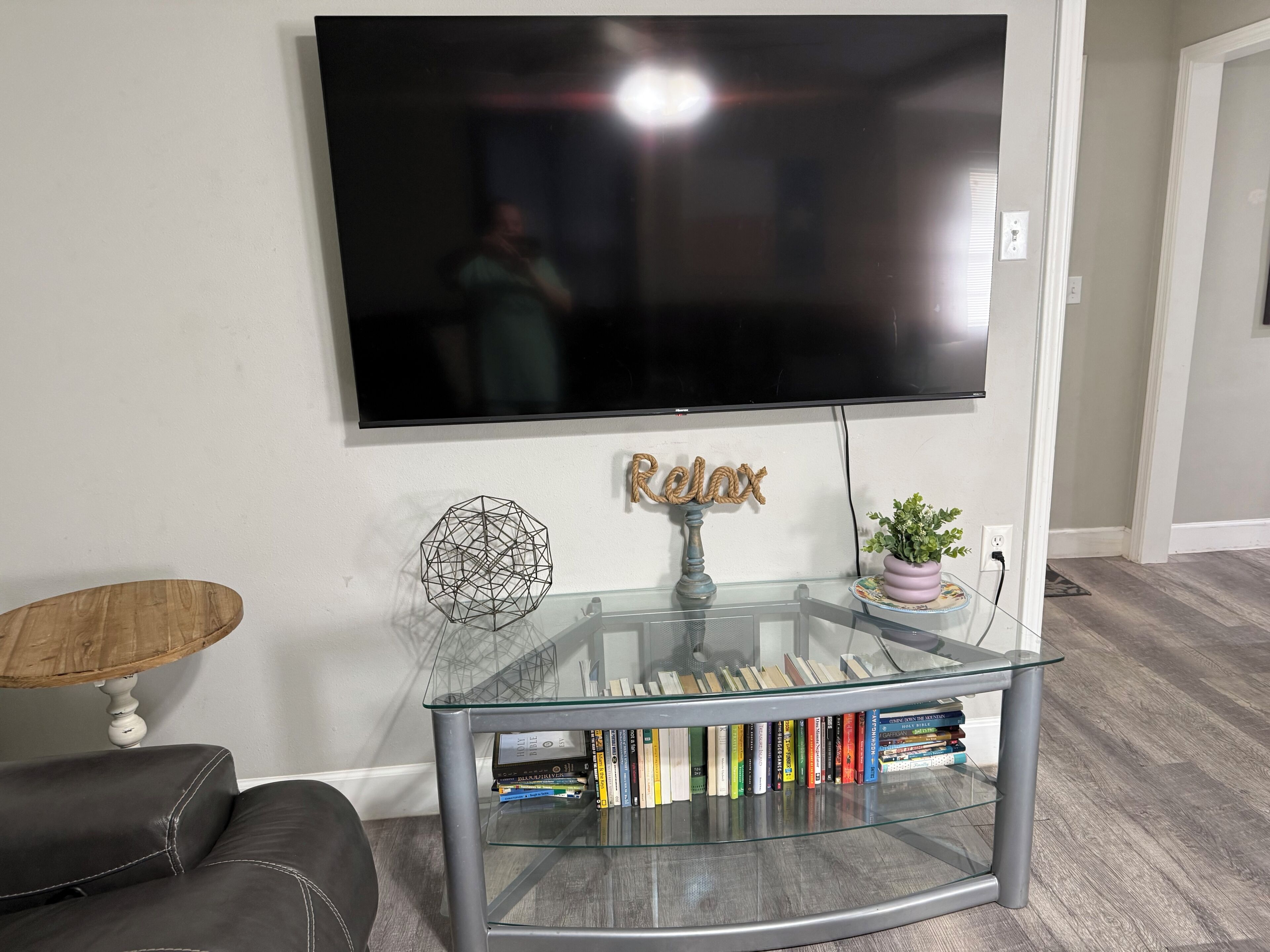 Een woonkamer met een flatscreen televisie aan de wand, een glazen salontafel met boeken, een decoratieve geometrische sculptuur en een bord met de tekst 'Relax'.