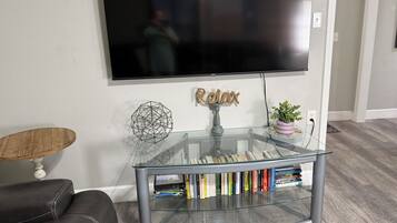 Een woonkamer met een flatscreen televisie aan de wand, een glazen salontafel met boeken, een decoratieve geometrische sculptuur en een bord met de tekst 'Relax'.