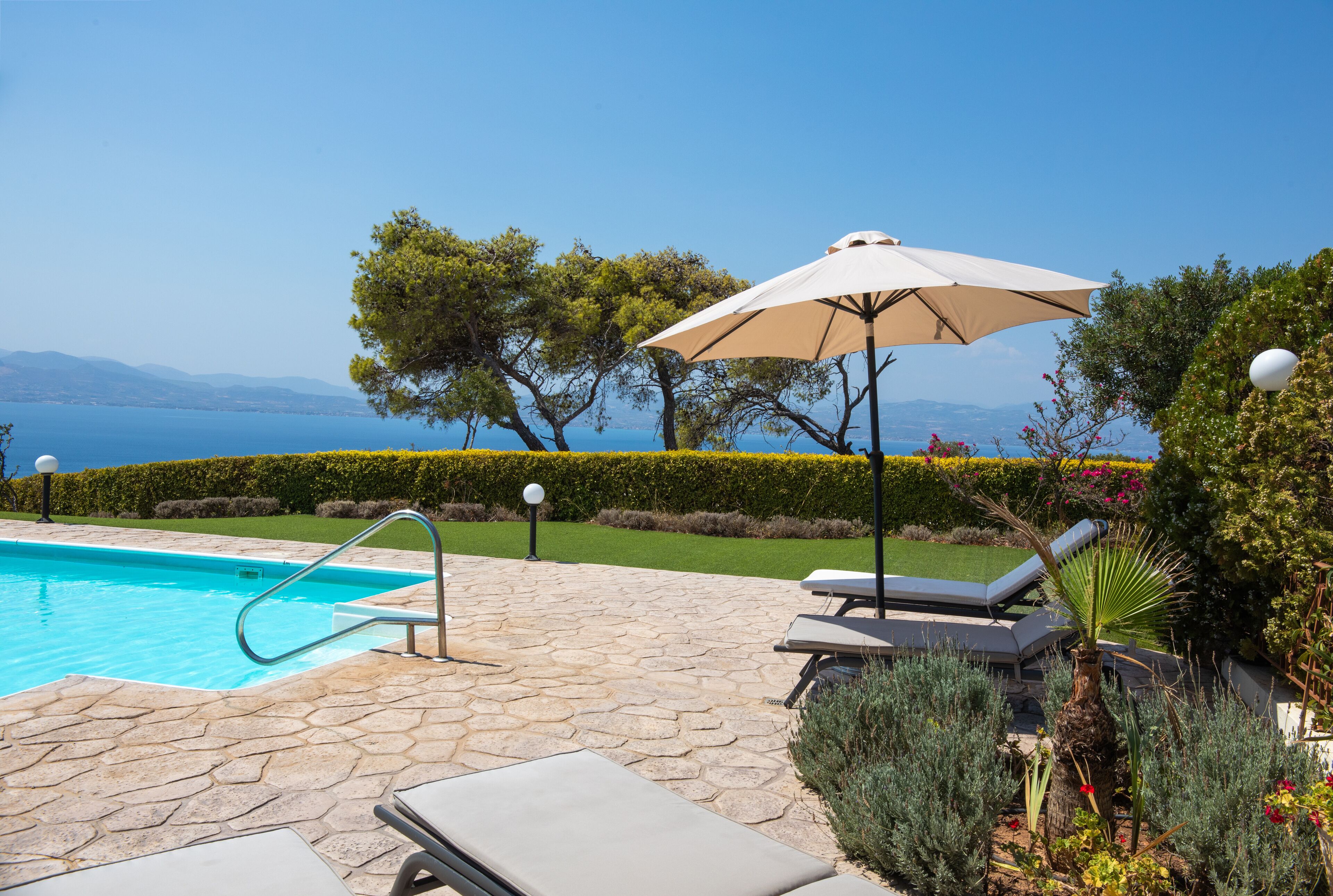 Ein Swimmingpool mit Liegestühlen und einem Sonnenschirm, umgeben von Grünflächen mit Blick aufs Meer.