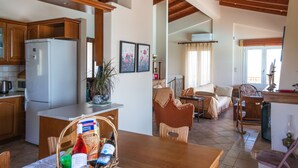 A spacious kitchen with wooden cabinets and a refrigerator, a dining area with a table and chairs, and a living room with a sofa and a window. - Lahtagas Estate with private swimming pool (Loutraki)