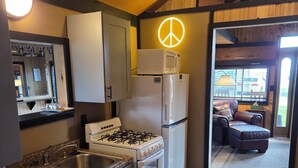 A compact kitchen with a gas stove, a microwave, and a refrigerator. There is a wooden ceiling and a view of an outdoor seating area through a glass door. - "That 70's Cottage" (on Lake Erie!) Free Firewood! (Essex)