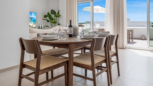 A dining area with a round table, four chairs, and a bottle of wine. There is a large window with a view of the sky and a plant in the corner.
