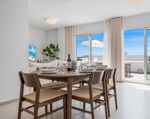 A dining area with a round table, four chairs, and a bottle of wine. There is a large window with a view of the sky and a plant in the corner. - Feliz | Roof terrace - pool - gym (Estepona)