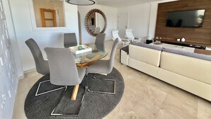 Ein modernes Wohnzimmer mit einem Glastisch, grauen Stühlen, einem weißen Sofa, einem an der Wand befestigten Fernseher und einem dekorativen Spiegel.