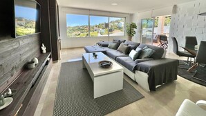 Ein modernes Wohnzimmer mit einem Ecksofa, einem Flachbildfernseher, einem Couchtisch und einem großen Fenster mit Blick ins Freie.