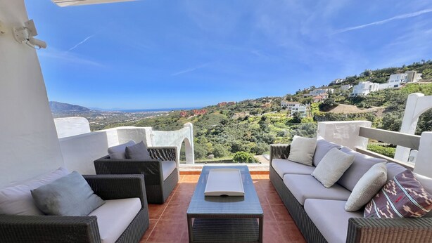 Eine Terrasse mit Korbmöbeln, einem Couchtisch und Blick auf eine Hügellandschaft mit Gebäuden.