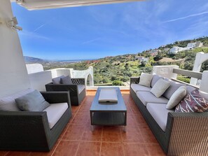 Une terrasse dotée de meubles en osier, d’une table basse et offrant une vue sur une colline parsemée de bâtiments.