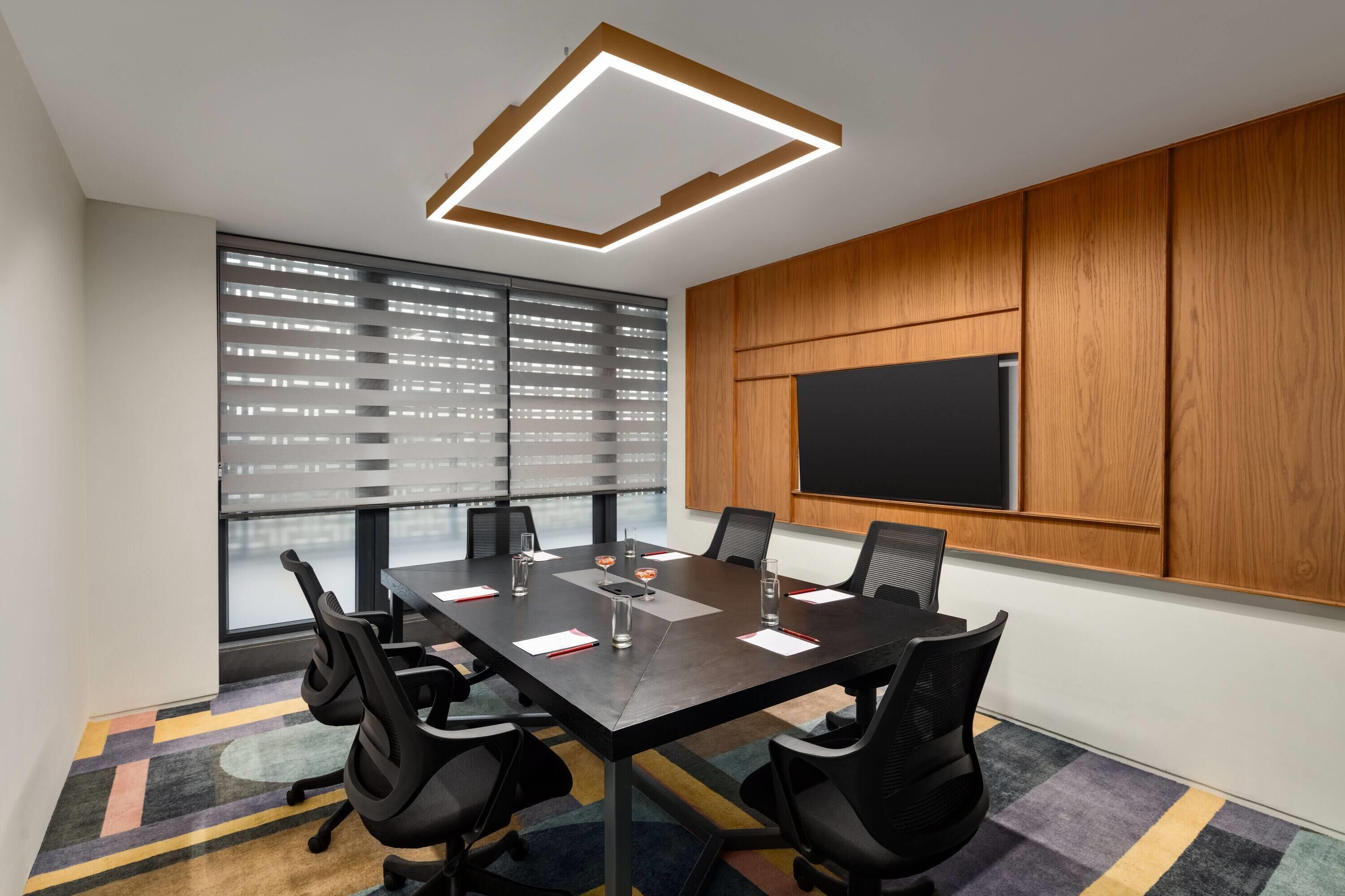 Une salle de conférence dotée d’une grande table, de chaises noires, d’un écran de projection et d’un mur recouvert de panneaux en bois.