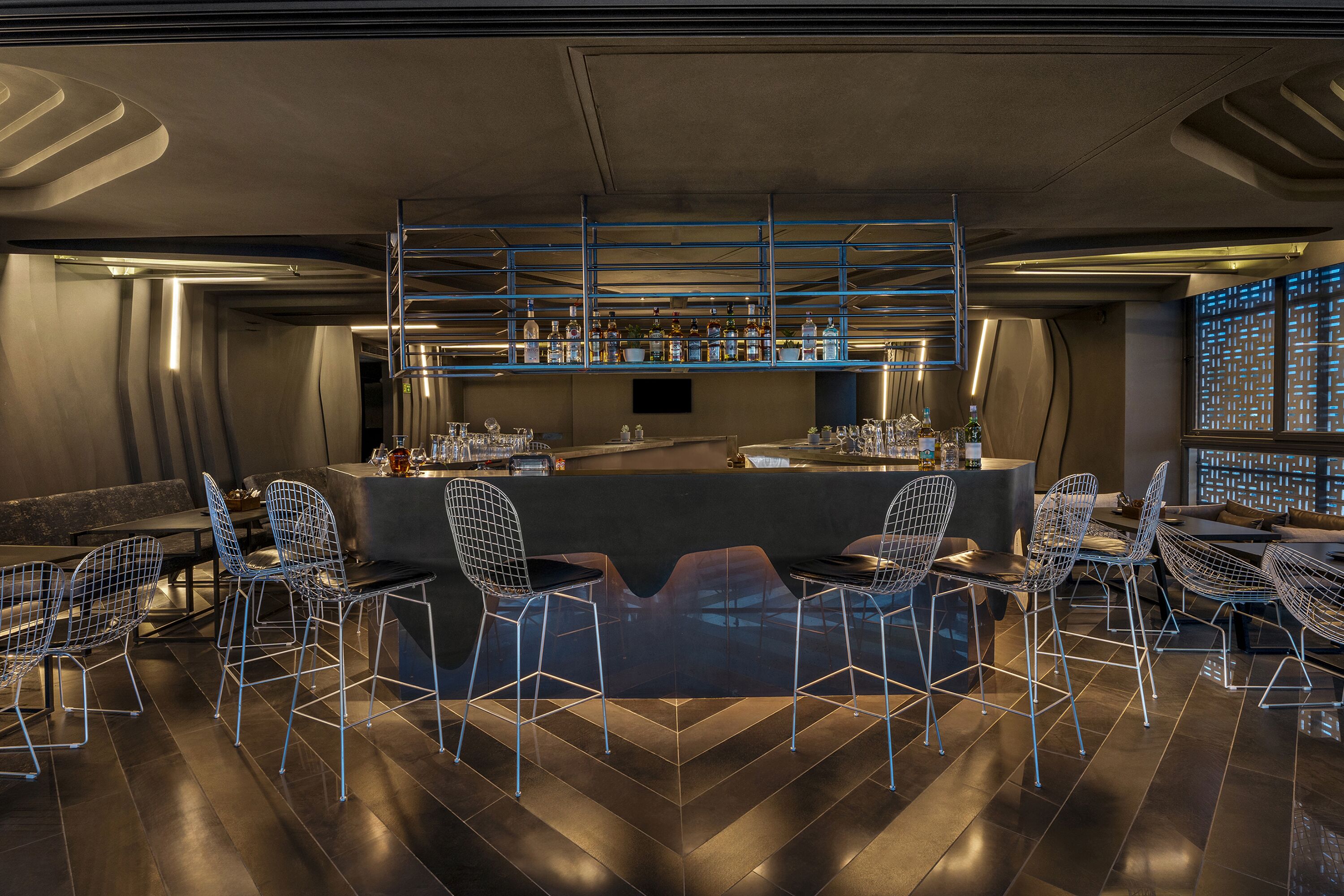 Eine moderne Bar mit Metallstühlen, einer Theke voller Flaschen und einem Fernsehbildschirm.