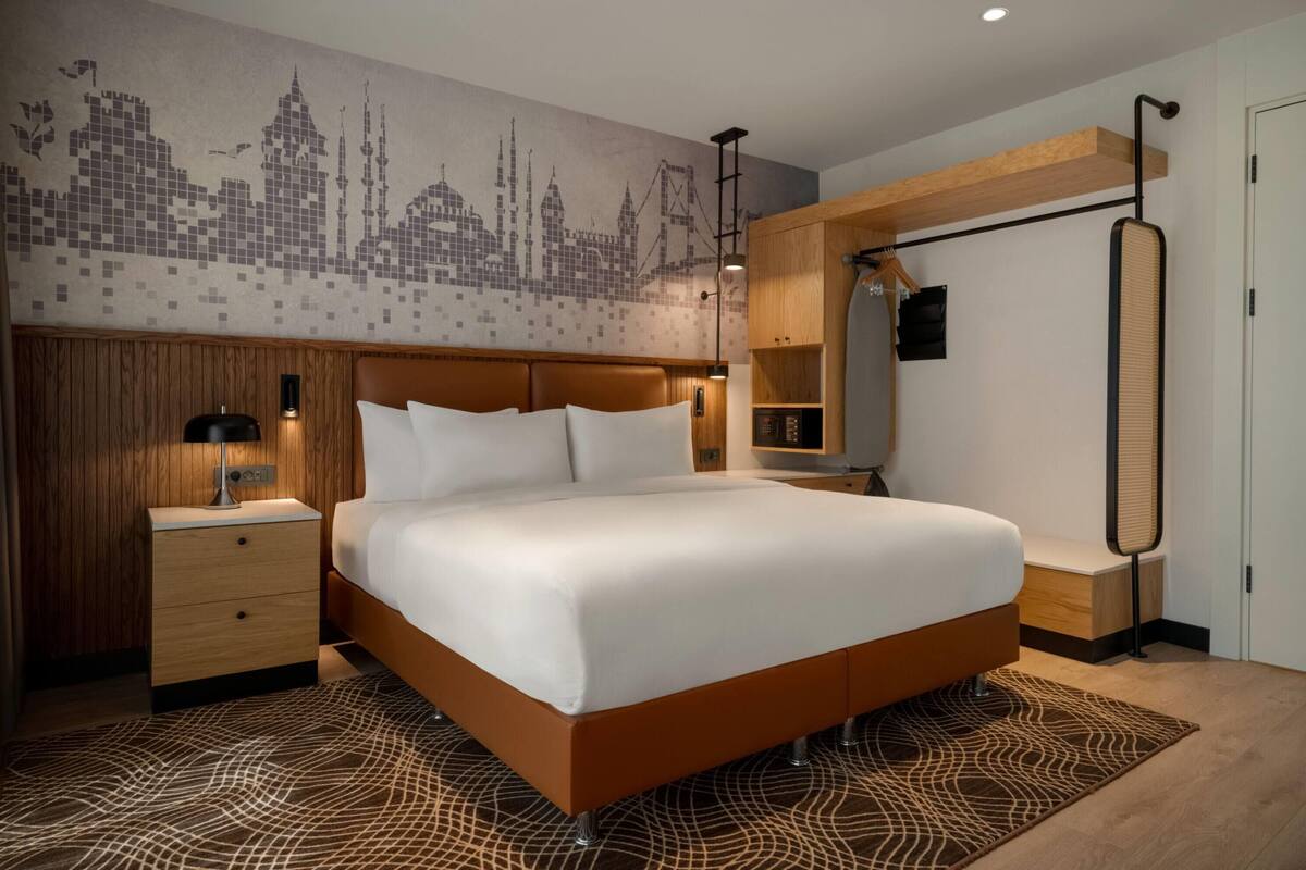 A modern hotel room with a large bed, wooden headboard, a cityscape mural, a hanging wardrobe, and a bedside table with a lamp.