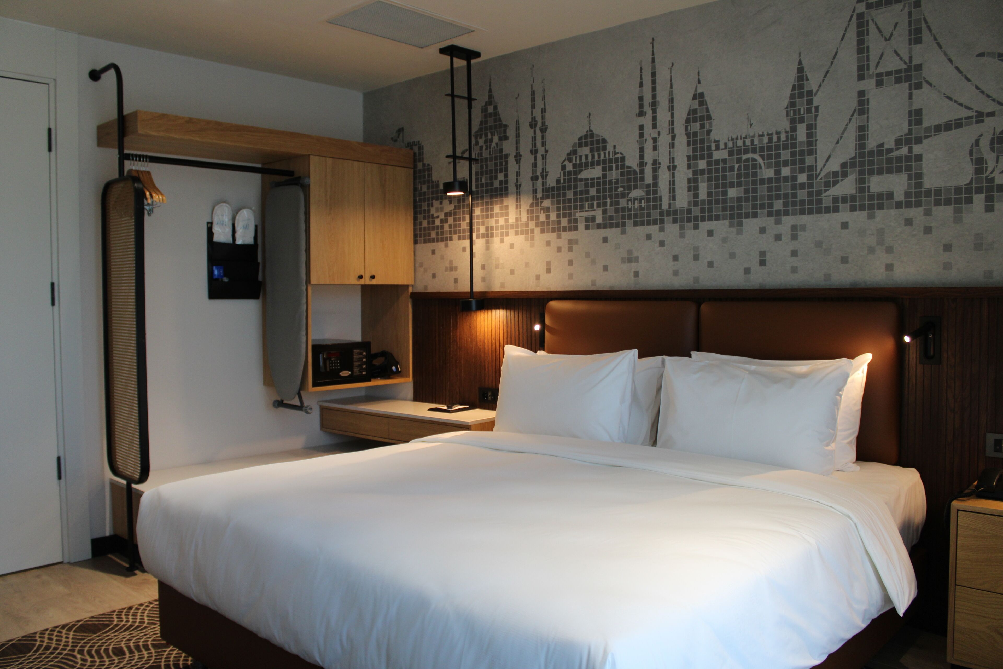 A hotel room with a large bed, a wooden headboard, a nightstand, a desk with a lamp, a wardrobe, and a wall with a cityscape mural.