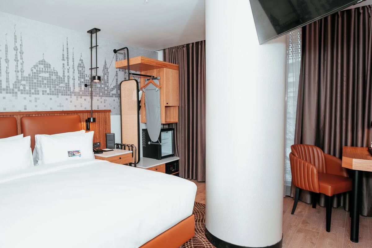 A hotel room with a large bed, a desk, a chair, a mirror, and a wall with a cityscape design.