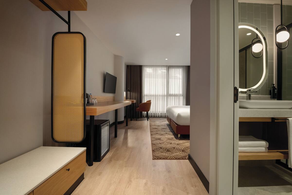 A modern hotel room with a wooden desk, a television, a bed with a patterned rug, and a bathroom with a large mirror and sink.