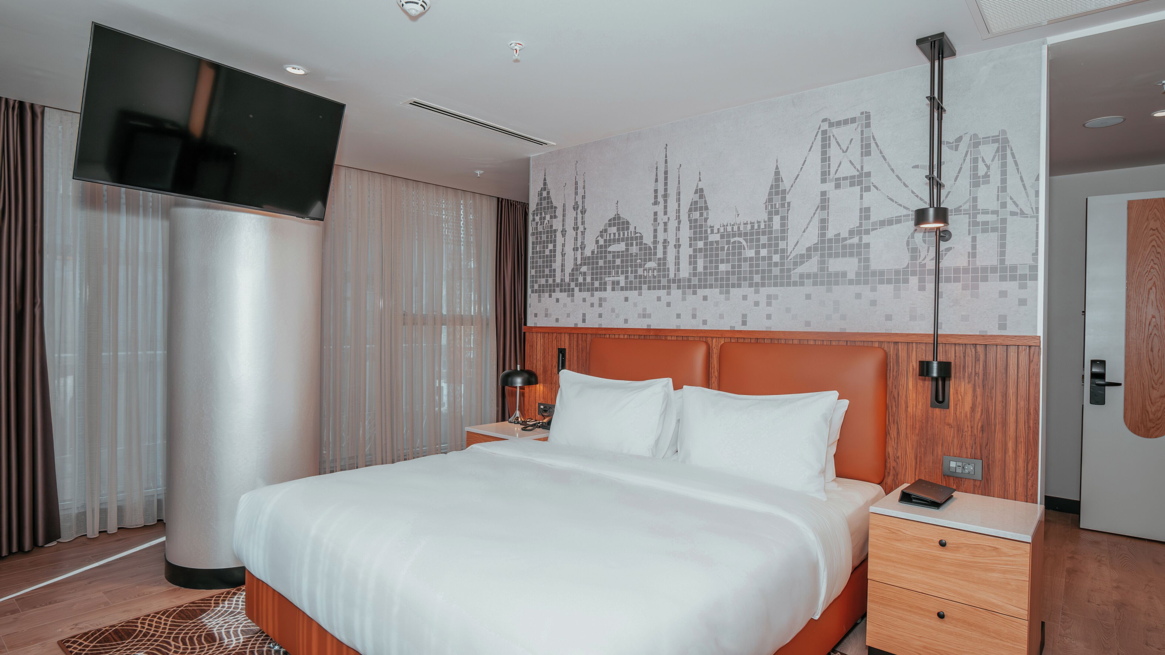 A hotel room with a bed, a TV, a nightstand, a lamp, and a cityscape mural on the wall.