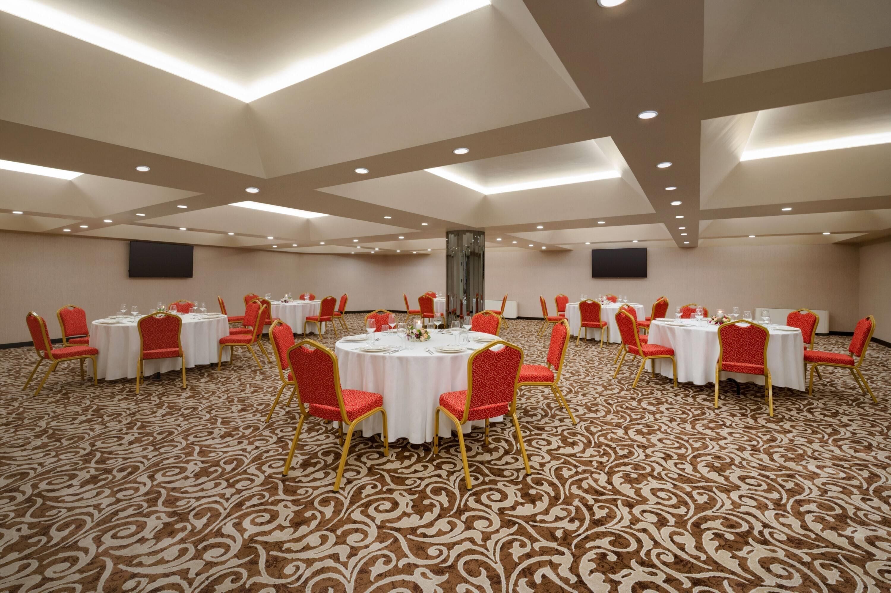 A large conference room with multiple round tables, each surrounded by red chairs and white tablecloths. The room has a patterned carpet and recessed lighting on the ceiling.