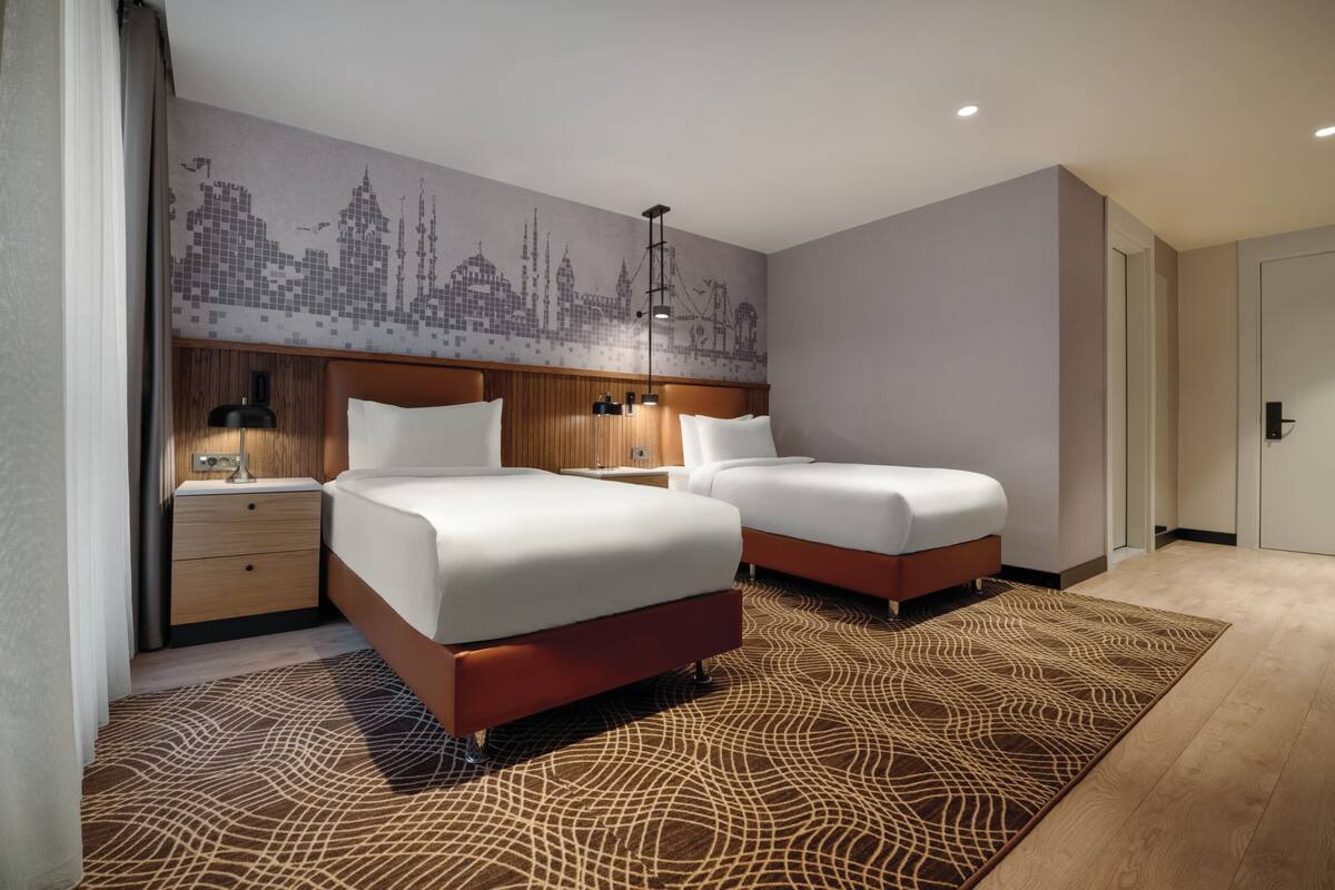 A hotel room with two beds, a cityscape mural, wooden headboard, nightstands, and a patterned rug.