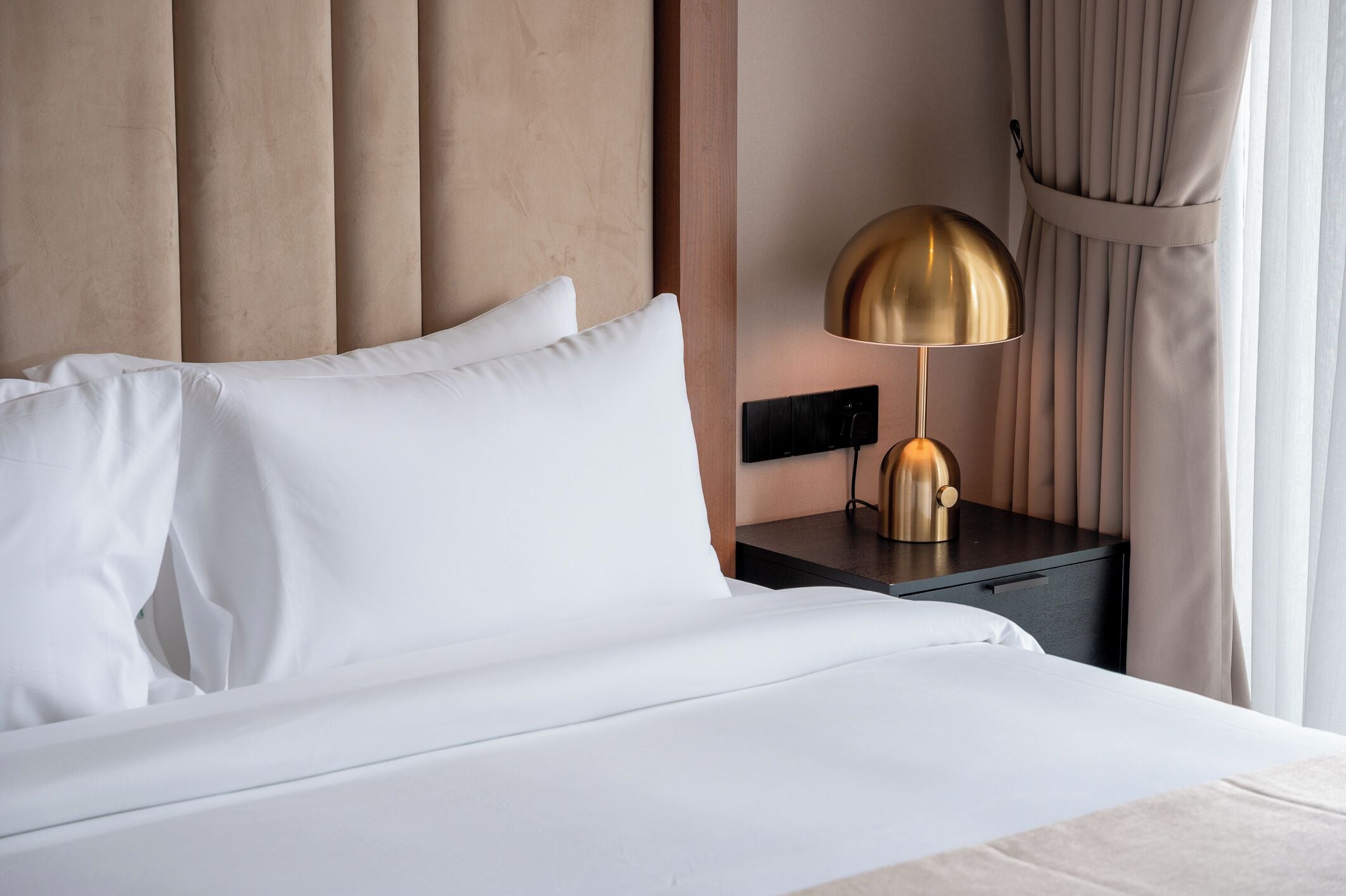 Una cama bien hecha con sábanas blancas, una lámpara dorada en la mesita de noche y una cortina en la ventana.
