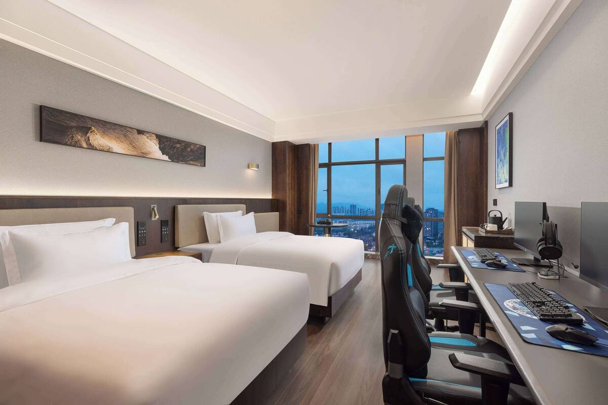 Ein Hotelzimmer mit zwei Betten, einem Schreibtisch mit Computer und einem großen Fenster mit Blick auf die Stadt.