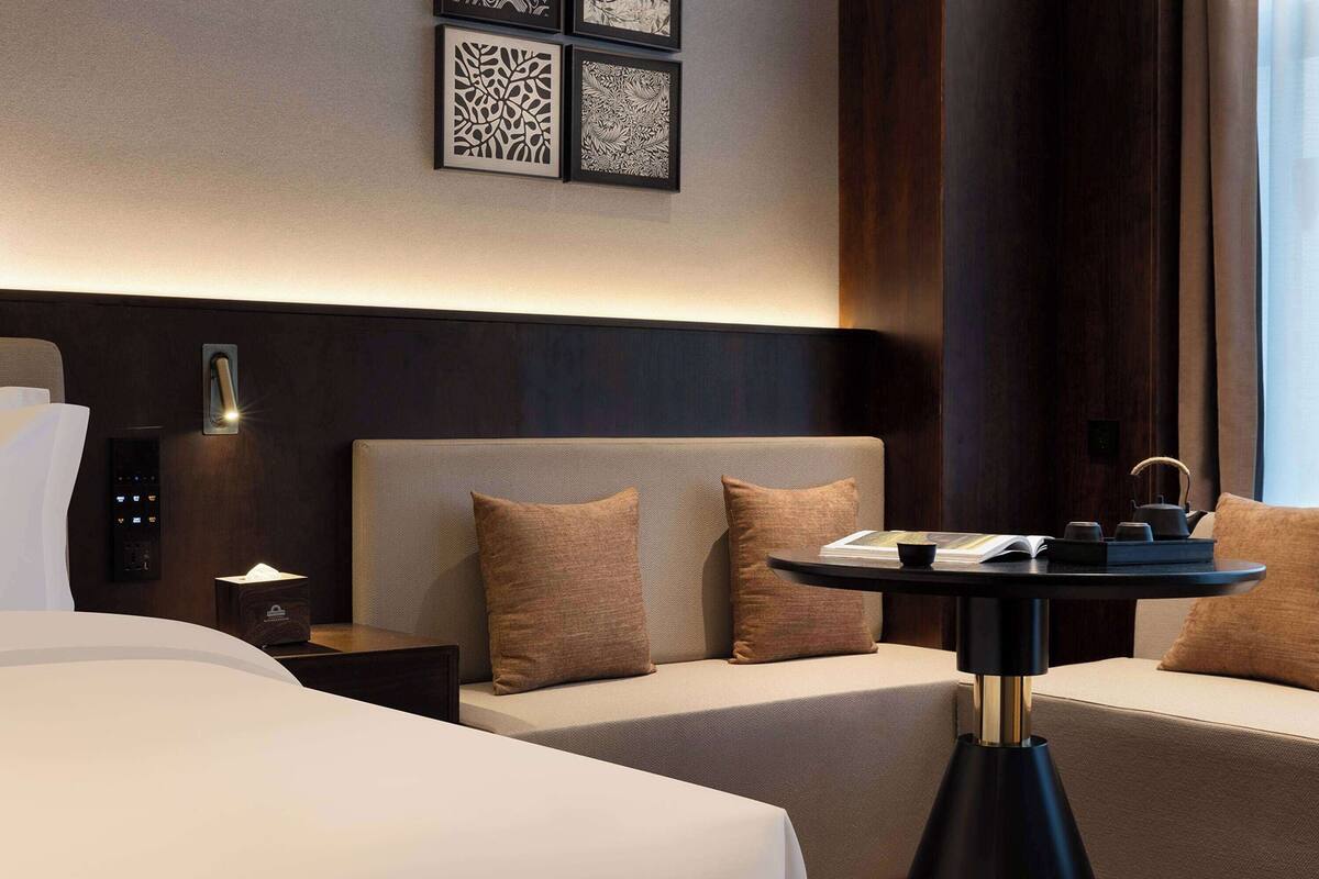 Ein Hotelzimmer mit einem Bett, einem Nachttisch mit Lampe, einem kleinen Tisch mit Tablett und einer Couch mit Kissen.