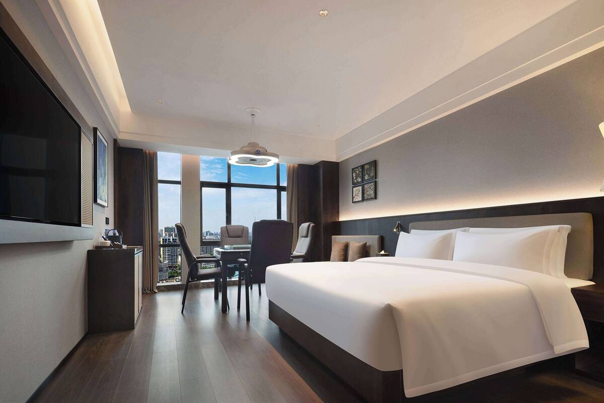 Ein modernes Hotelzimmer mit einem großen Bett, einem Schreibtisch mit Stuhl, einem Fernseher und Blick auf die Stadt durch große Fenster.