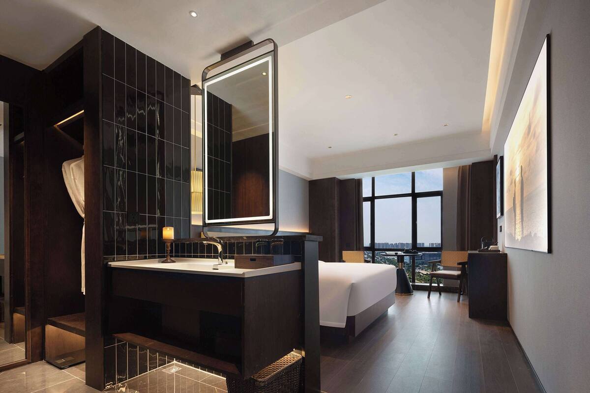 Ein modernes Hotelzimmer mit einem großen Spiegel, einem Bett, einem Schreibtisch und Blick auf die Stadt durch große Fenster.