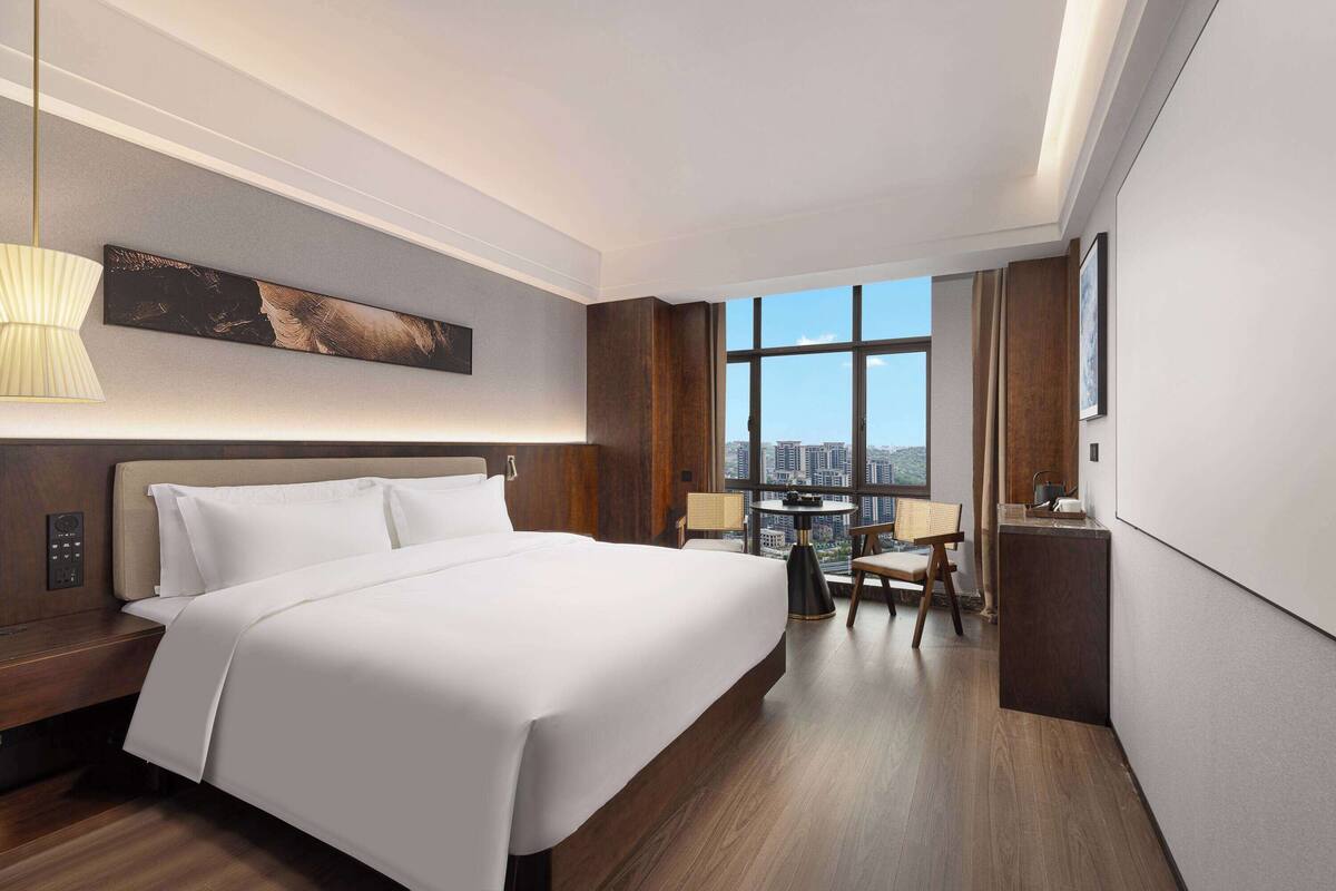 Ein modernes Hotelzimmer mit einem großen Bett, einem Schreibtisch und Blick auf die Stadt durch ein großes Fenster.