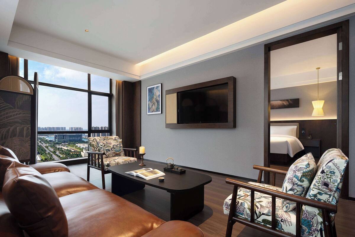 Ein modernes Hotelzimmer mit einer Ledersofa, einem Couchtisch, gemusterten Sesseln, einem Flachbildfernseher und einem großen Fenster mit Blick auf die Stadt.