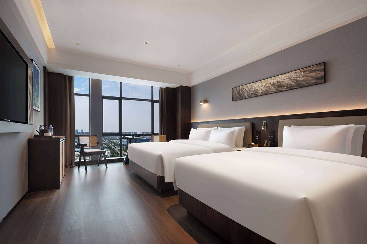 Ein Hotelzimmer mit zwei Betten, einem Schreibtisch, einem Stuhl und einem großen Fenster mit Blick auf die Stadt.