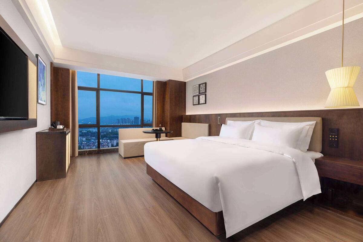 Ein modernes Hotelzimmer mit einem großen Bett, einem Fernseher, einem Schreibtisch und Blick auf die Stadt durch große Fenster.